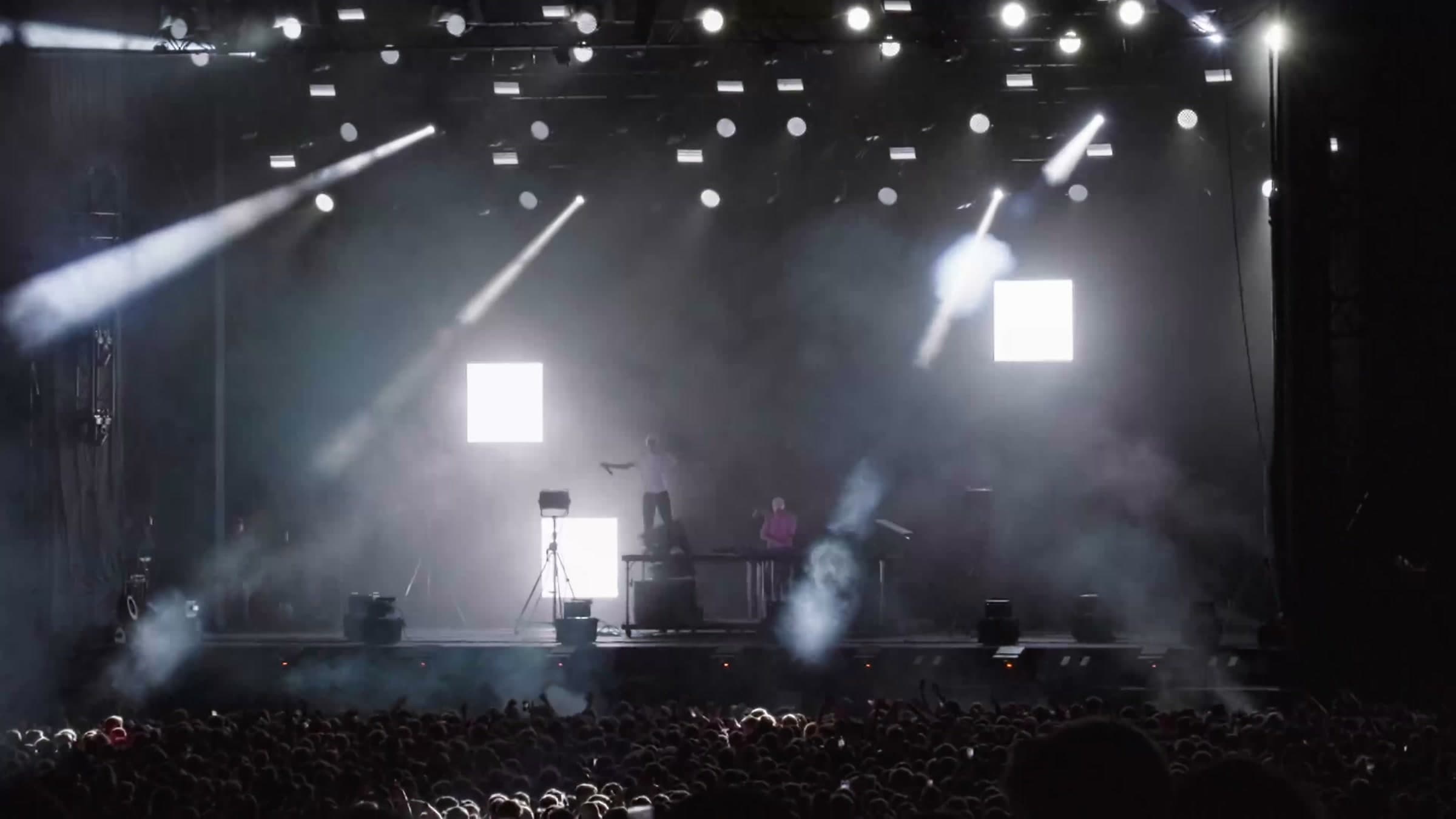 Scene med kryssende lysstråler og silhuettert publikum under en konsertkveld på Festningen