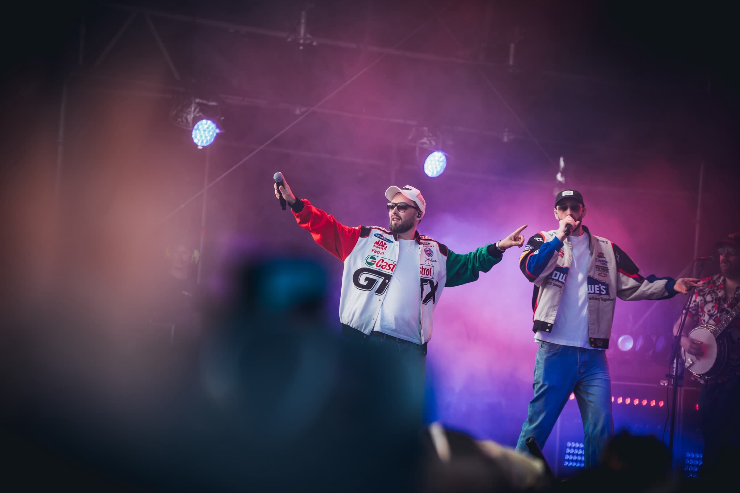 To artister i fargerike jakker opptrer på en festivalscene med lilla scenelys