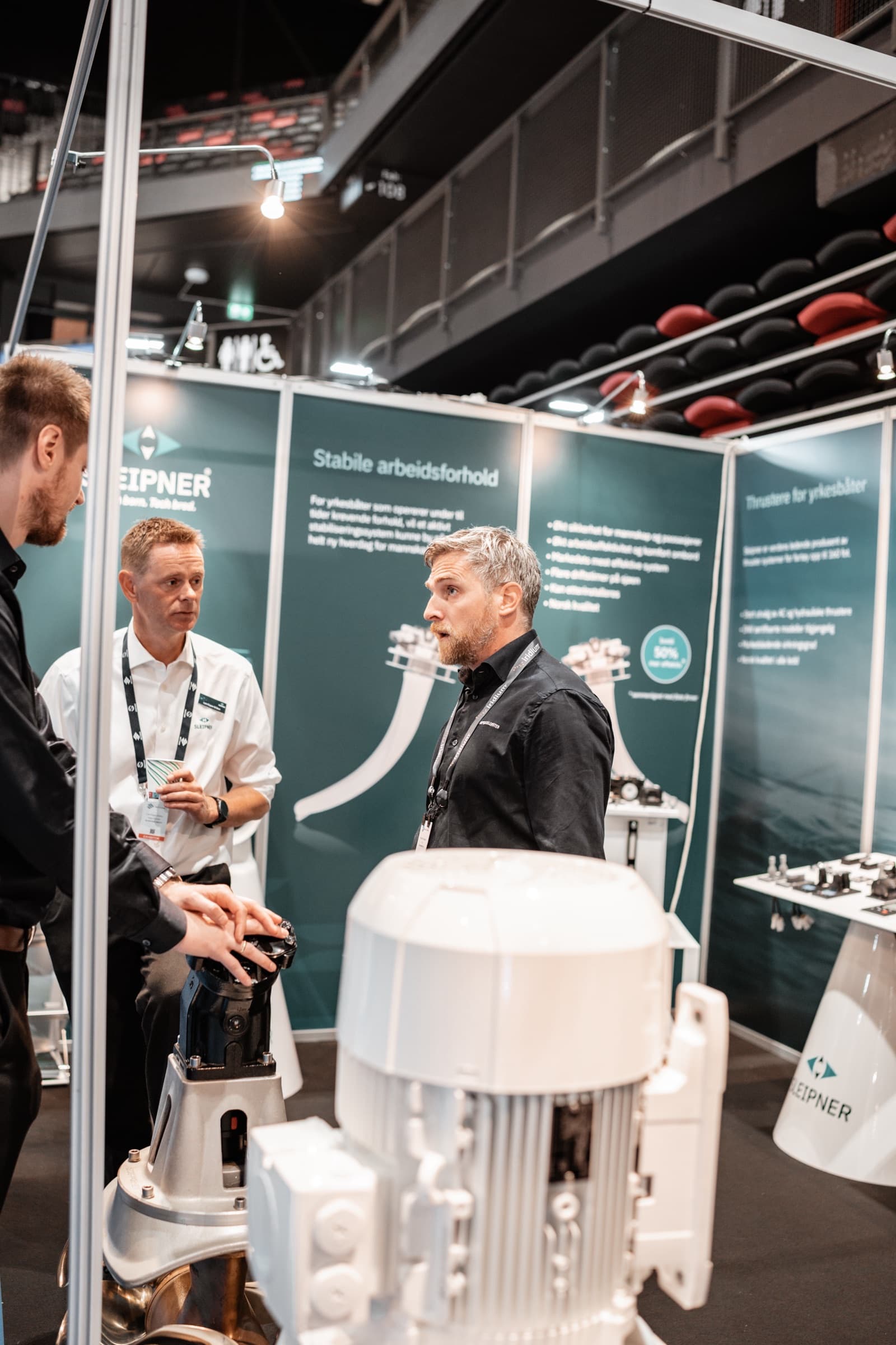 Selgere i samtale med besøkende ved en messestand med skipsteknisk utstyr